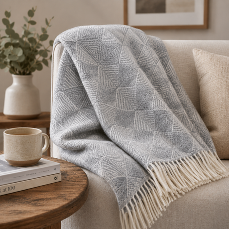 A grey wool blanket with a diamond pattern draped over a beige sofa beside a side table with books and a coffee mug
