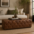 A brown tufted leather ottoman in a styled living room setting, positioned in front of a cream sofa and decorated with a tray, book, and a small potted plant.