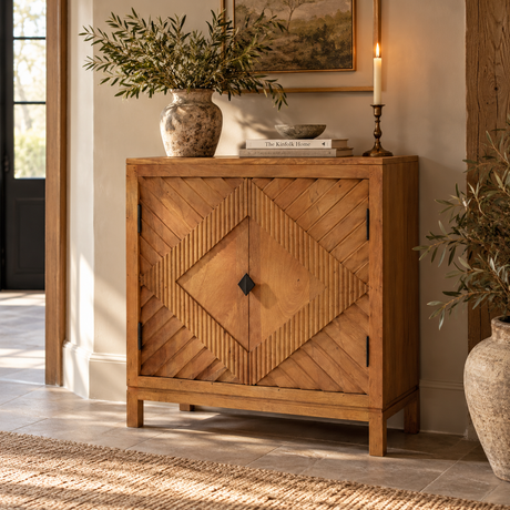 A rustic wooden sideboard cabinet with geometric carved panel doors, styled in a warm, sunlit room next to a vase of greenery and a candle.