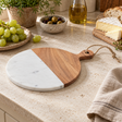 The Loxley Round Marble and Acacia Wood Paddle Board with Acacia Handle on a stone kitchen worktop beside grapes, olives, bread and cheese.