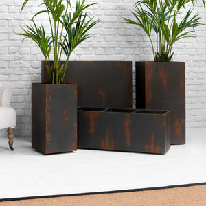 A set of four rectangular metal planters with a distressed rust finish, arranged on a white floor against a white brick wall, with two green palm plants placed inside them.