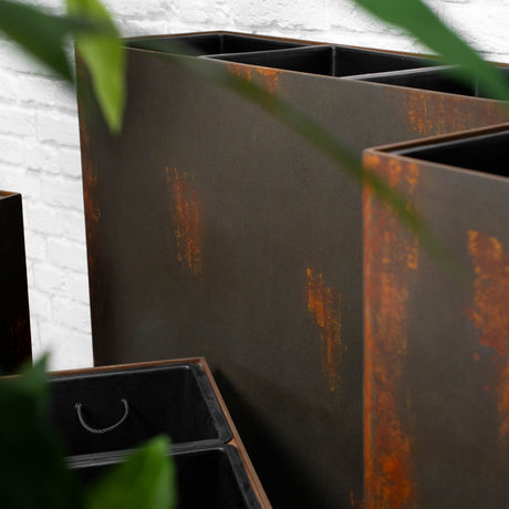 Rectangular metal planter boxes with a dark charcoal finish and rusted orange-brown weathered accents, shown in an outdoor setting with green leaves in the foreground.