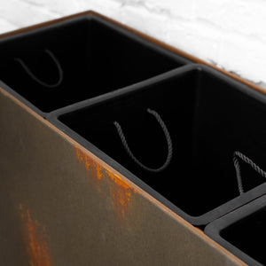A close-up of a rectangular metal container with three dark compartments, featuring a rusted brown-orange finish on the side and black interiors with twisted rope handles visible inside.