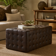 A dark brown tufted leather ottoman in a cozy living room setting, styled with books and a decorative leafy branch on top.