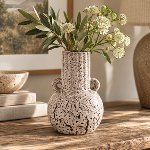 Freya Speckled Brown Stoneware Bud Vase with a flared neck and two loop handles, filled with green leaves and white flowers, set on a rustic wooden table in a warm, styled interior.