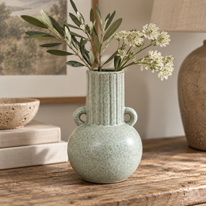 Freya Speckled Green Stoneware Bud Vase with a fluted neck and two loop handles, holding olive branches and small white flowers, styled on a wooden table with books and a lamp in a warm indoor setting.
