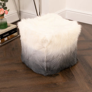 Fluffy white to grey ottoman on a wooden floor with books and a plant in the background.