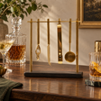 Loxley bar tool set with black slate and gold stand on a wooden sideboard beside decanters and cocktails