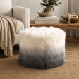 A round goatskin ottoman with a white-to-grey ombre gradient, styled in a cosy living room setting next to a beige armchair and a wooden side table.
