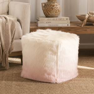 A square faux-fur ottoman stool in a soft white-to-pale-pink ombre gradient, styled in a cozy living room setting next to a beige armchair and a wooden side table.