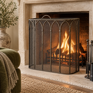 Gothic antique pewter fire screen in front of a lit open fire in a cream stone fireplace, beside a green velvet armchair.