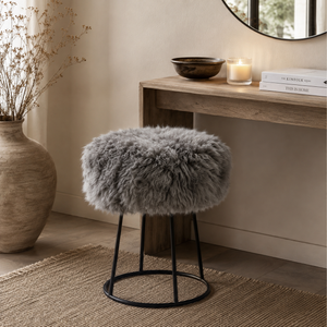 A round grey sheepskin stool with a black metal wire frame, styled in a warm, neutral-toned room next to a wooden console table.