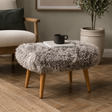 A round sheepskin ottoman stool with light wood legs in a styled living room setting, featuring a grey and white shaggy texture, wooden legs, and a small black mug on top.