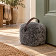 Grey sheepskin doorstop with leather handle holding open a sage green door on a stone hallway floor