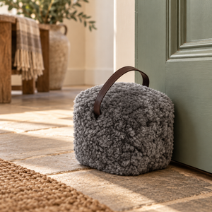 Grey sheepskin doorstop with leather handle holding open a sage green door on a stone hallway floor