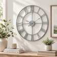 Large decorative grey skeleton wall clock with Roman numeral markers on a wall with plants and books below.