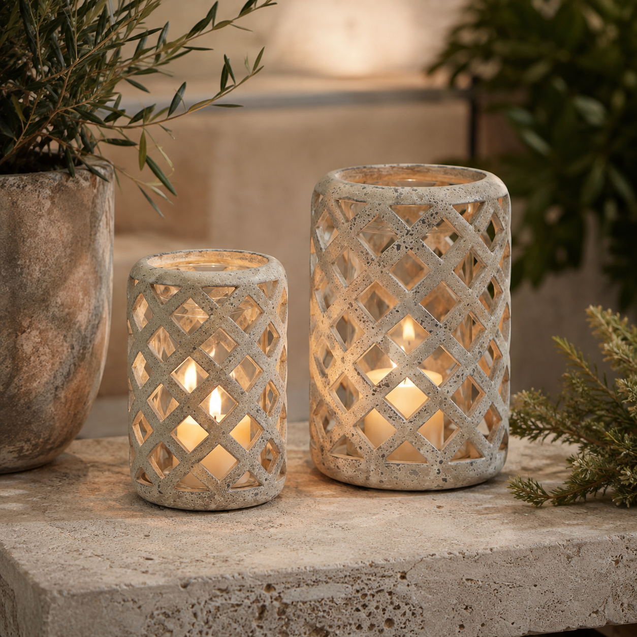 Two decorative candle holders with a distressed stone-look lattice pattern, one small and one large, each containing a lit tea light candle, displayed on a stone surface with plants in the background.