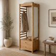 A tall light oak wood open wardrobe with a built-in mirror, two drawers at the bottom, and a coat hanging on a hanger. The coat rack is styled in a warm, minimalist room with a potted plant, a side table, and neutral-coloured walls.