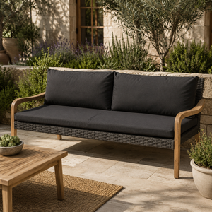 Outdoor patio sofa with black cushions and a woven wicker-style frame, shown in a garden courtyard setting with a wooden table and plants.