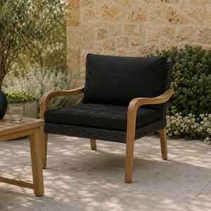 A natural lifestyle image of a wooden outdoor lounge chair with black cushions and dark woven detailing, styled on a sunlit stone patio surrounded by greenery.