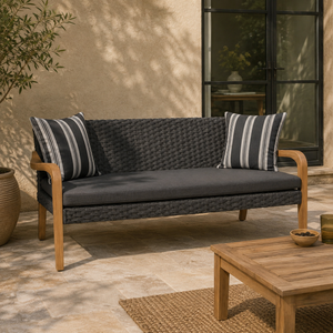 Outdoor patio loveseat with a dark woven wicker-style seat and back, light natural wood arms and legs, a dark grey cushion, and two striped throw pillows in a sunlit patio setting.
