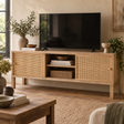 A light natural wood TV stand with woven cane doors and open shelving, styled in a living room setting with a black television, decorative books, and plants.
