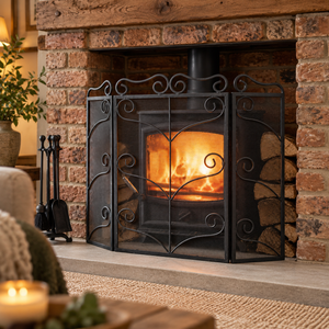 Black ornate three-panel fire screen in front of a lit wood burner in a brick fireplace, with logs stacked beside it.