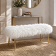 A white faux fur bench with slim gold metal legs, styled in a bright entryway next to a console table.
