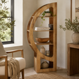 A tall, curved wooden bookshelf with open shelves stands in a styled room corner beside a window, holding books, bowls, vases, and small plants. A cream upholstered chair with a beige throw is partly visible on the left, and a wooden side table with a plant is on the right.