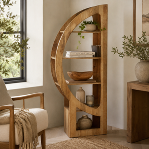 A tall, curved wooden bookshelf with open shelves stands in a styled room corner beside a window, holding books, bowls, vases, and small plants. A cream upholstered chair with a beige throw is partly visible on the left, and a wooden side table with a plant is on the right.