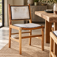 The Jakarta Cowhide and Teak Wood Dining Chair with a cream hair-on cowhide seat and back, at a rustic wooden dining table on a jute rug.
