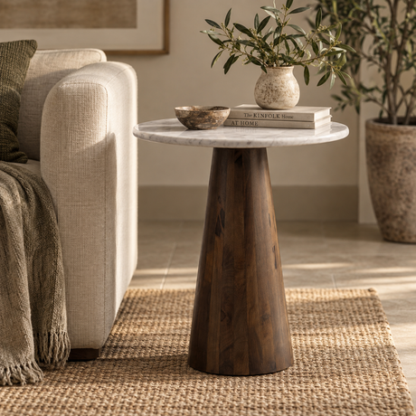 A dark brown wooden side table with a round white marble top, styled in a cosy living room next to a beige sofa with a throw blanket.