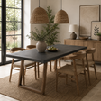 A modern dining room scene featuring a dark gray rectangular concrete table with light wood legs, surrounded by woven and wood dining chairs on a jute rug. The room is styled with wicker pendant lights, a wooden sideboard, and a large potted plant.