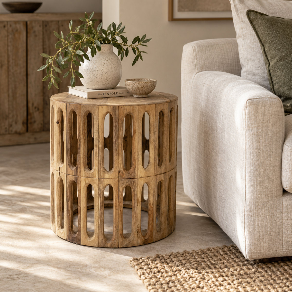 A round wooden side table with open cutout panels, styled in a living room next to a light beige armchair with a vase and small bowl on top.