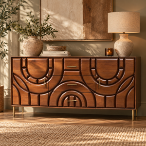 A mid-century modern wooden sideboard with carved geometric patterns on the front, styled in a warm, sunlit room with a lamp, potted plant, and wall art.