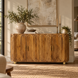 A mid-century modern wooden sideboard with a carved arched panel design, styled in a warm, sunlit living room with a large ceramic vase, candles, and wall art.