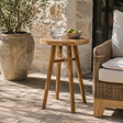 A round wooden side table with three legs, styled on a stone patio next to a wicker armchair and a large potted plant.
