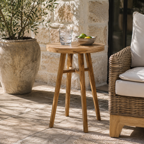 A round wooden side table with three legs, styled on a stone patio next to a wicker armchair and a large potted plant.
