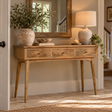 A rustic wooden console table with three drawers and tapered legs, styled in a warm entryway with a mirror, lamp, ceramic vase, and plants.