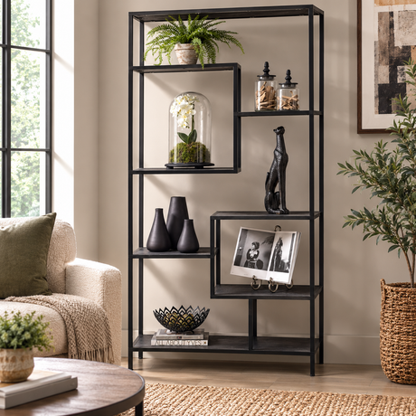 A black metal open shelving unit with six tiers stands in a styled living room setting, holding various decorative items including plants, vases, a small sculpture, and a framed photo.