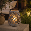 A decorative outdoor candle holder with a textured stone-look finish and a diamond lattice cutout pattern, containing a warm glowing candle. It is placed on a stone ledge in a garden setting at night.