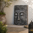 A rectangular stone Buddha head wall sculpture with a dark grey textured finish, displayed in a garden setting against a light beige wall with pebbles, a potted plant, and a decorative lantern.