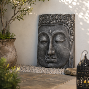 A rectangular stone Buddha head wall sculpture with a dark grey textured finish, displayed in a garden setting against a light beige wall with pebbles, a potted plant, and a decorative lantern.