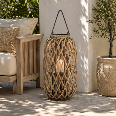 A tall cylindrical woven rattan lantern with a black hanging rope and a lit candle inside, placed on a patio beside a wooden chair and a potted plant.