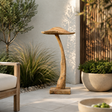 A mushroom-shaped wooden ornament made from natural tree trunk and branches, placed on a light stone patio in a garden setting with plants and a chair nearby.