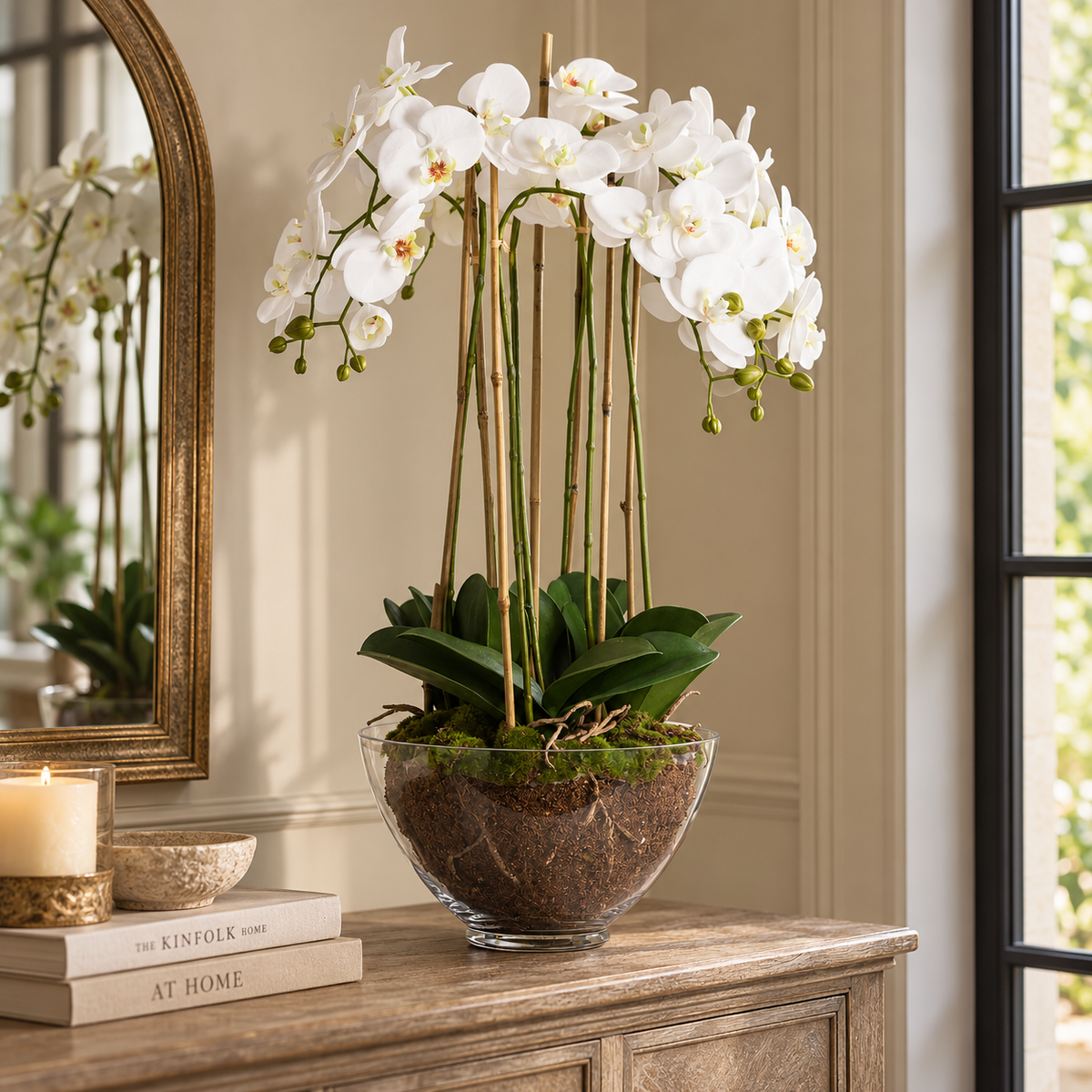 Large White Orchid In Glass Pot on a wooden sideboard beside an arched gilt mirror, a lit candle, a stone bowl and books