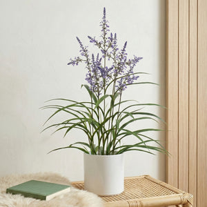 Artificial lavender plant with delicate purple flowers and green foliage in a white pot, styled on a natural rattan tray indoors.