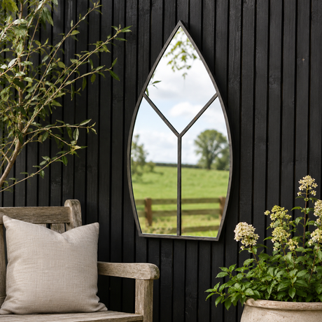 A decorative arched mirror with a dark metal frame hangs on a black wood-paneled wall. The mirror reflects a green field under a cloudy sky, while the surrounding scene includes a wooden bench with a beige pillow on the left and a potted plant on the right.