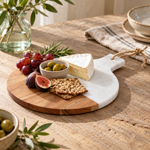 The Loxley Round Marble and Acacia Wood Paddle Board with Marble Handle, with cheese, figs, grapes, olives and crackers on a rustic wooden table.