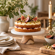 The Loxley Round White Marble and Acacia Wood Cake Stand holding a layered fig and pistachio cake on a rustic wooden table with candles and flowers.